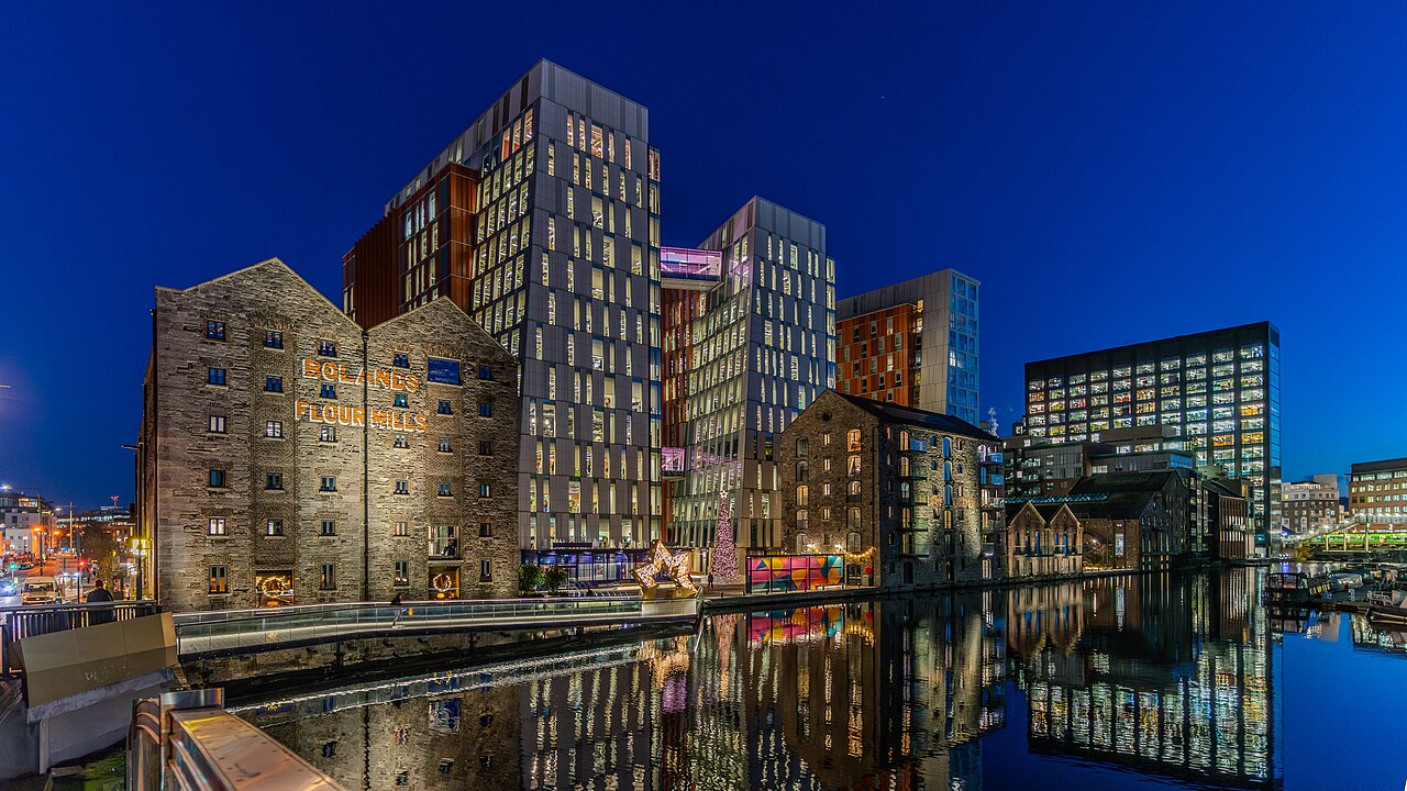 A wide-angle night photograph of the Boland's Quay redevelopment in Dublin, Ireland. The image features the illuminated historic stone facade of the "Bolands Flour Mills" on the left, contrasted with modern glass-and-steel high-rise office buildings in the center. The buildings are reflected in the calm waters of the Grand Canal Dock in the foreground. A illuminated Christmas tree and star are visible in the plaza.
