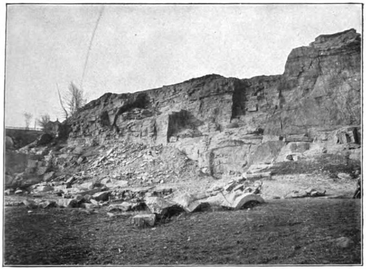 Plate IV A Original caption:  "Gaither's Granite Quarry, Ellicott City, Maryland."
Text from the volume referring to the figure:
Several quarries opened in the steep and moderately high granite cliffs along the east side of Patapsco River have been operated nearly opposite Ellicott City. The largest of these, known as the Gaither quarry, comprises two large openings that adjoin each other and have been worked back from the river into the granite cliffs, with an elevation from the base to the top of about 75 feet. (See Pl. IV, A.)
The rock is a biotite granite of medium dark blue-gray color and medium grain. Its structure is decidedly foliated or schistose, though much less pronouncedly so than that of the granite gneiss at Port Deposit and Frenchtown; and its original granite characters are still apparent. Hand specimens of the granite show abundant fine granules of yellow epidote closely associated with the biotite. Irregular grains of brownish allanite are also apparent in the hand specimens. Because of its richness in epidote and allanite Keyes classified this granite and some of the granites from the Port Deposit, Woodstock, Dorseys Run, and Ilchester localities as allanite-epidotebearing granites.

The granite consists of soda-lime feldspar (oligoclase), potash feldspars (orthoclase and microcline), quartz, and biotite, together with accessory zircon, apatite, hornblende, allanite, epidote, and secondary chlorite and epidote. Intergrowths of the feldspar and quartz in more or less rounded grains are of common occurrence. The feldspar is partly cloudy and opaque from alteration, and the larger individuals inclose small rounded grains of feldspar and quartz. Allanite and epidote occur as parallel growths. Partial granulation of the quartz and feldspar into fine mosaics from pressure metamorphism is pronounced.