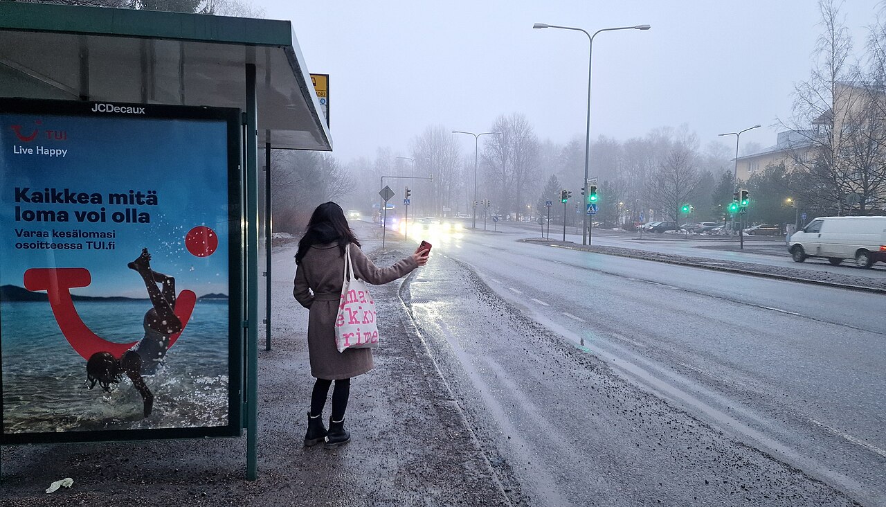 Matkustaja näyttämässä pysähtymismerkkiä bussille Linnanrakentajantiellä: Sumuinen ja harmaa kevättalven näkymä bussipysäkille, jossa on aamuseitsemän aikaan matkustaja. Taustalla Laajasalon suunnasta saapuva bussi. Etualalla pysäkin seinässä oleva TUI:n matkailumainos. Oikealla Kaivolahdenkatu ja ohitse ajava valkoinen pakettiauto.