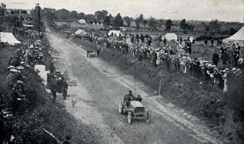 1903 Gordon Bennett Trophy. René de Knyff, driving his Panhard to second place, passes Alexander Winton repairing the Winton Bullet 2 on the first lap.