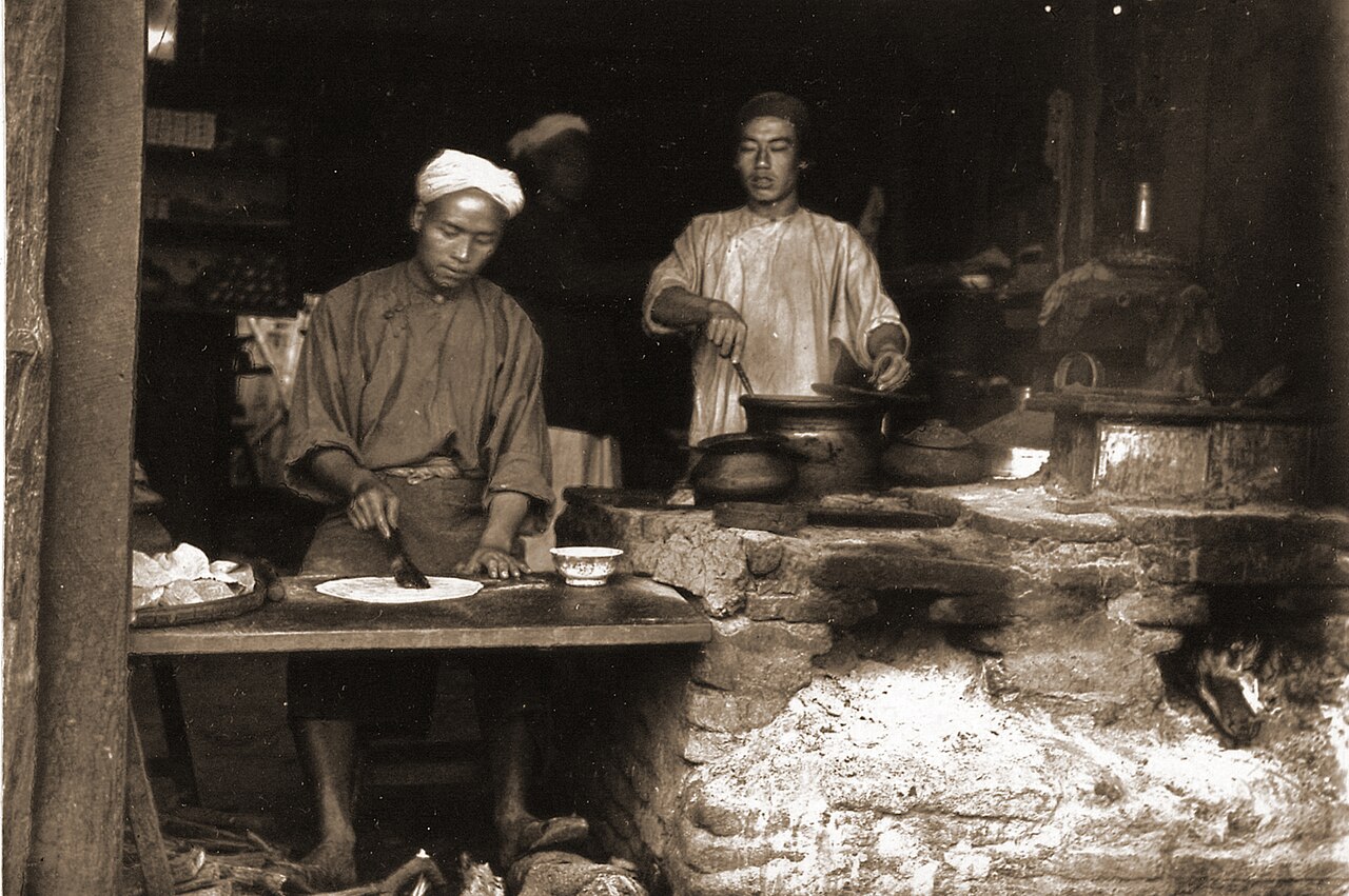 Photograph of a baker’s shop in the Chinese quarter of Mandalay in Burma (Myanmar), taken by Willoughby Wallace Hooper in 1886.
 The war culminated in the annexation of Upper Burma by the British on 1 January 1886. Chinese communities had existed for many centuries in rural Burma and were largely formed by migrants travelling overland from China into Burma along the north-eastern trade routes. The urban Chinese population originated during the colonial era when Chinese from the coastal provinces of China came by sea to work as merchants, among other professions. The Chinese quarter in Mandalay was near the King’s Bazaar, on the south side of B Road. A caption by Hooper accompanying the photograph describes the shop: “It is not perhaps a very inviting looking place, nevertheless the bread made in Mandalay is exceedingly good. Bread is much eaten by the Burmese, besides various kinds of cakes and chupatties, a sort of pancake or girdlecake: the man on the left is making one of these while the other man is tending the pot over the furnace.”