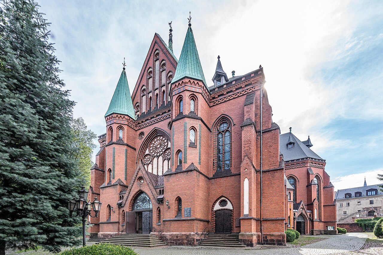 Saint Nicholas Church in Nowa Ruda, at 9 Kościelna Street in Nowa Ruda, Kłodzko County, Lower Silesian Voivodeship, Poland, on 6 July 2024. The photograph shows a close view of the Gothic Revival brick parish church from the façade and side elevation, with a massive body, tall pointed-arch windows, rose window, ceramic decoration, buttresses, portals, greenish tower roofs and spires. The frame also includes entrance steps, a paved church square, lamps, greenery and trees. The image documents historic sacred architecture of Nowa Ruda, one of the important Gothic Revival churches of the Kłodzko Land and an object entered in the register of monuments.