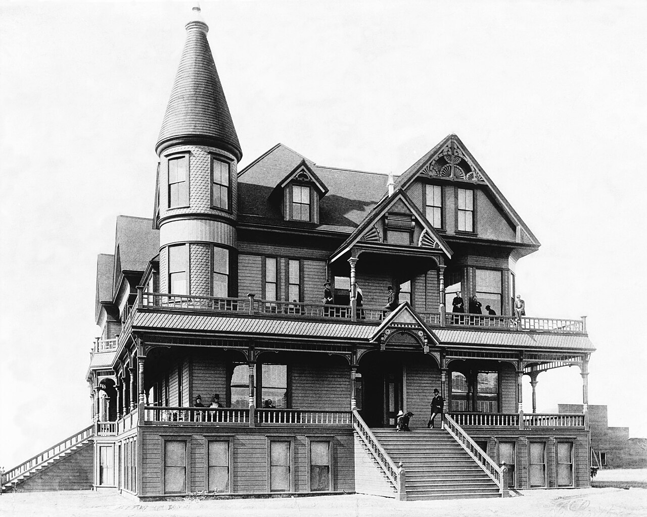 Photograph of an exterior view of the Mission Hotel in San Fernando, eastern San Fernando Valley ca.1888 (1874?). 
• The three and a half-story, Victorian-style hotel is shown at center, with people posing on its porch and balcony.
• On the second floor a man can be seen leaning against one of the beams which support the overhang near the hotel's entrance. A dog is visible near him at the top of the front steps that lead to the second floor and the subsequent entrance.
• On the second floor to the left, a two-story tower extends from the hotel, decorated in gingerbread clapboard.