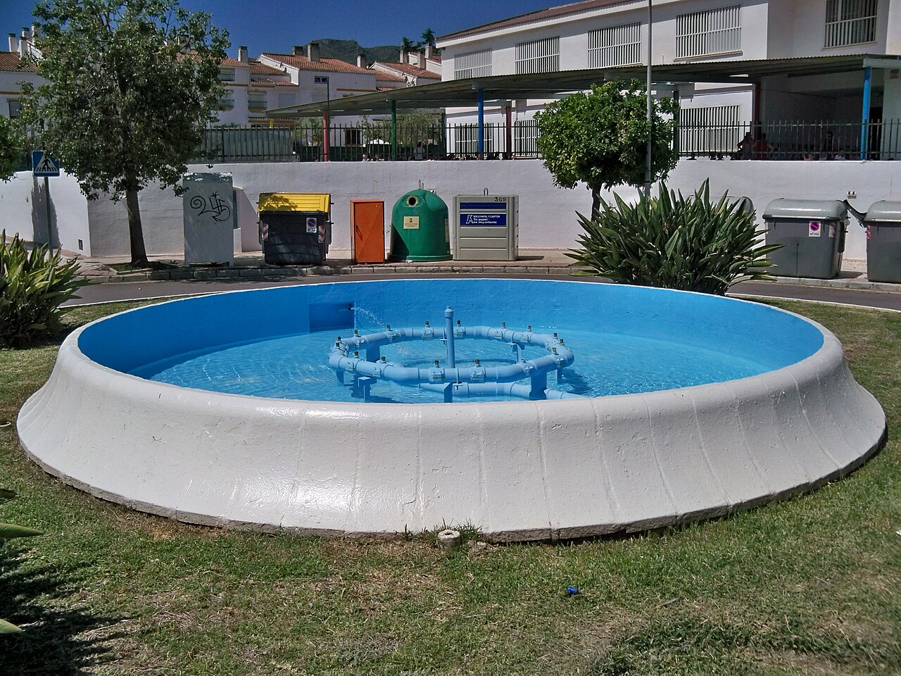Fuente de Sedella, en la calle Sedella, barrio Heliomar, distrito Churriana, Málaga, España. La fuente se encuentra en una pequeña rotonda de la calle. Consta de un estanque circular formado por bloques de hormigón de perfil curvo pintados de blanco y un aro de surtidores cuyos chorros forman una palmera.[1]