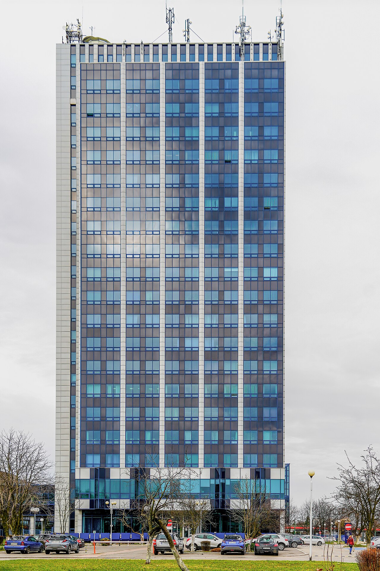 Building of the Faculty of Natural Sciences of the University of Silesia, commonly called “Żyleta”, at 60 Będzińska Street in Sosnowiec, Pogoń district, Silesian Voivodeship, Poland, on 22 February 2024. The photograph shows the south-eastern side of the high-rise building: a slender modernist volume, a repetitive rhythm of blue and brown glazing, vertical white façade strips, the lower part with entrance area, car park and greenery, antennas and telecommunication equipment on the roof, and a grey winter sky. The object is the tallest building in Sosnowiec and one of the most recognizable buildings of the University of Silesia; it houses the Faculty of Natural Sciences of the University of Silesia, formerly the Faculty of Earth Sciences, and the Museum of Earth Sciences.