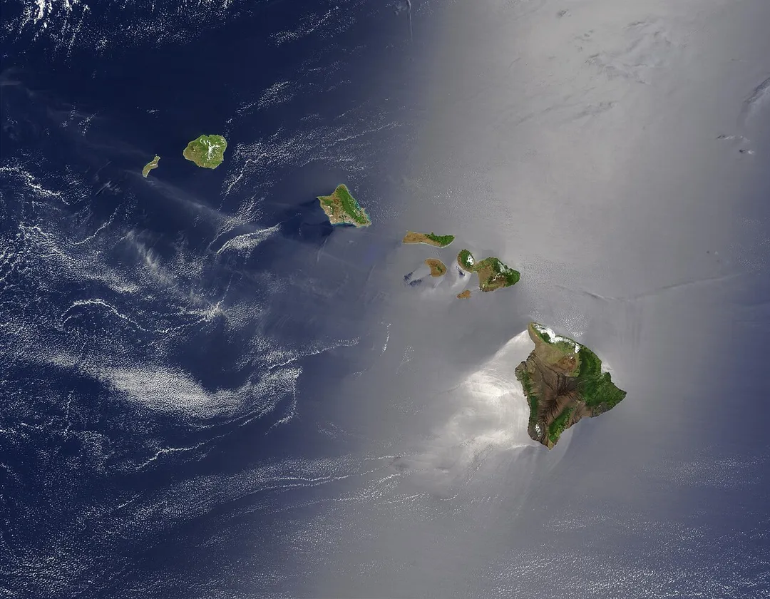 Satellite view of Hawaii archipelago (USA)  Original description from NASA:

"A silver swath of sunglint surrounds half of the Hawaiian islands in this true-color Terra MODIS image acquired on May 27, 2003. Sunglint reveals turbulence in the surface waters of the Pacific Ocean. If the surface of the water was as smooth as a perfect mirror, we would see the circle of the Sun as a perfect reflection. But because the surface of the water is ruffled with waves, each wave acts like a mirror and the Sun’s reflection gets softened into a broader silver swath, called the sunglint region.
"In this scene, the winds ruffling the water surface around the Hawaiian Islands create varying patterns, leaving some areas calmer than others. Southwest of Hawaii and Maui, on their leeward sides, calmer waters are indicated by brighter silver coloration. Conversely, notice how most of the vegetation on the Hawaiian Islands grows on their northeastern, or windward, sides.

"From lower right to upper left, the “Big Island” (Hawaii), Maui, Kahoolawe, Lanai, Molokai, Oahu, Kauai, and Niihau islands all make up the state of Hawaii, which lies more than 2,000 miles from any other part of the United States."