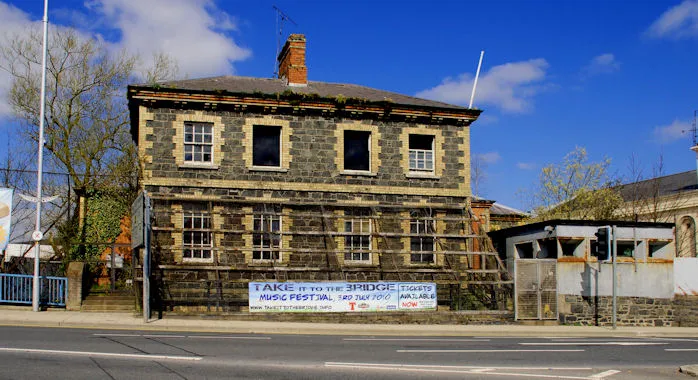 Former police station, Banbridge Built circa 1860 as the Downshire estates office and later acquired as an RUC barracks.  Now replaced by a police station nearby, it lies forlorn and empty in Church Square, still with all the embellishments of The Troubles.  During the 1956/62 campaign it merely had a few sandbags against which the constabulary parked their bicycles.  On the At Risk register.