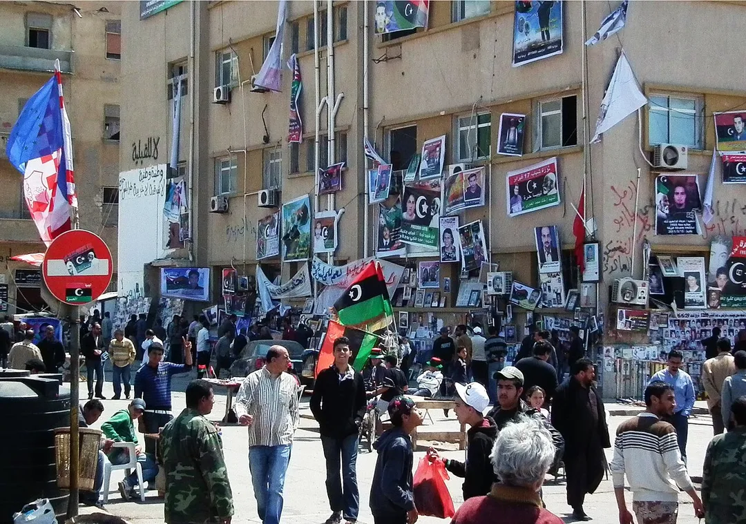 Crowd at the Court house square in Benghazi, April 2011. The square was a place for gathering, demonstrations and organisation of the uprise movement. The walls of the court house are draped with pictures of casualties of the fighting, as well as former executions by Gaddafi, mourners passing by. Flags and souvenirs are sold at a table in the center. The red-white flag on the left marks the al-Ahly soccer club that was destroyed by Gaddafi's order in 2000, and three leaders executed, after insubordination.