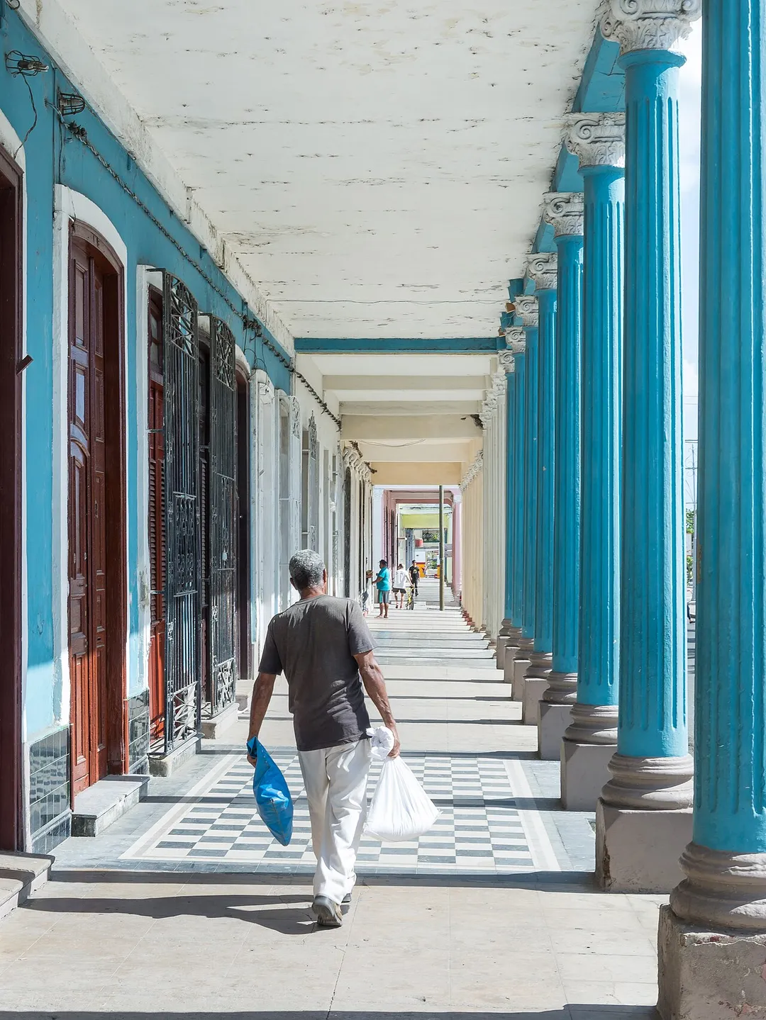 Man carrying a heavy bag through a seemingly endless colonial style corridor in Cienfuegos, Cuba.