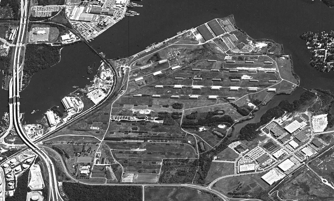 Oblique view of Curtis Bay Depot, Defense Logistics Agency site, in 1984.  Ordnance Road is along the bottom, and north is to the left.  The highway along the left edge is I-695, the Baltimore Beltway.  The channel in upper left is Curtis Creek, and the stream at right is Back Creek.  Many of the buildings adjacent to Ordnance Road have been demolished to create a sports park with multiple baseball fields.  Most other buildings on the site are in disrepair and are collapsing (as of 2021), but some close to the riverfront are still in use.