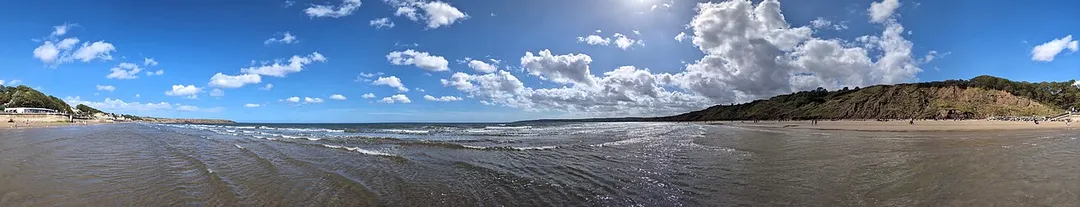 This panoramic photograph, captured on 31st August 2025, shows Muston Sands in Filey, North Yorkshire. The vantage point, from the water's edge near the foot of Martin's Ravine Slipway, offers a 360 degree view of the coastline, and a classic British seaside scene.
The composition is dominated by a vast, brilliant blue sky, characteristic of a fine late summer's day. It is filled with an array of distinct cloud formations. 
In the foreground, the shallow waters of the North Sea gently wash over the sand. The tide was on its way out, and the wide, wet shoreline mirrors the bright sky.
Distinct features of the Filey coastline frame the scene. To the right, a prominent stretch of low, earthy-brown boulder clay cliffs rises above the beach. These cliffs are topped with a layer of green vegetation and trees contrast with the golden sand below. The beach on this side is dotted with numerous people, appearing as small figures enjoying the sunshine, walking along the water's edge, or sitting in scattered groups.
To the far left, the panorama looks back towards the town of Filey. A man-made breakwater juts out into the sea, marking the southern end of the main promenade. Beyond this, the distant silhouette of the coastline stretching towards Filey Brigg is faintly visible.

Overall, the photograph encapsulates a perfect moment at the British coast. The panorama emphasising the immense feeling of open space, from the wide arc of the sky to the broad sweep of the bay, capturing the light, atmosphere, and gentle holiday activity of a beautiful day in North Yorkshire.