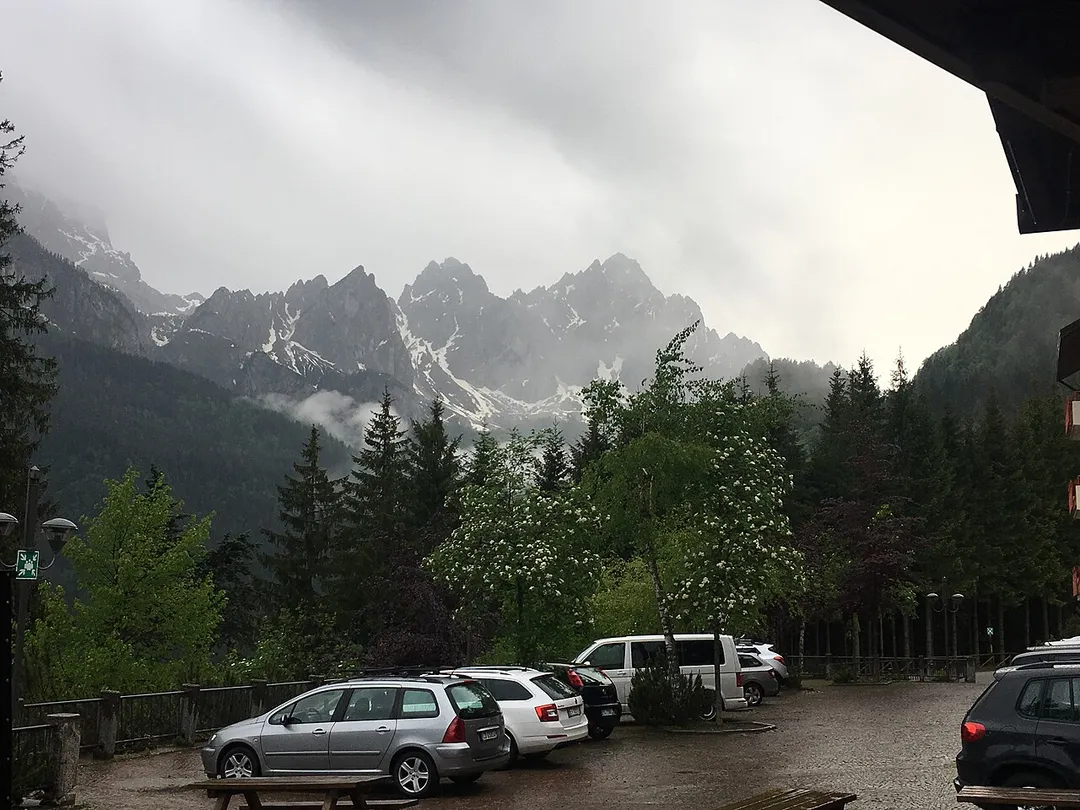 Photo from parking log, cars in the foreground and the beautiful mountains of Forni Avoltri in the background, the peaks covered in snow