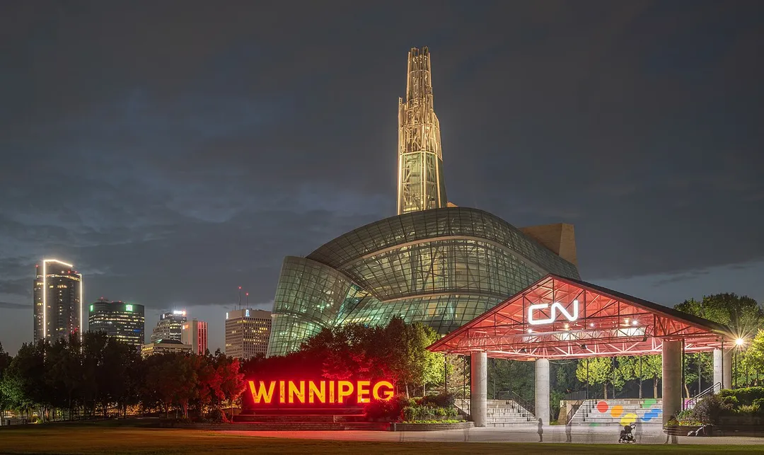 The Canadian Museum for Human Rights at night, viewed from The Forks in Winnipeg, Manitoba, Canada, with its illuminated glass atrium and spire rising above the downtown skyline. In the foreground, the illuminated "WINNIPEG" sign and the CN pavilion are visible, with blurred figures from a long exposure adding a sense of movement to the scene.