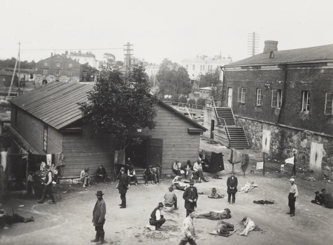 Red Guard prisoners of war at the Susisaari prison camp of Suomenlinna Fortress' IV district, the yard of the Adlerfeldt traverse, Adlerfeldt tarverse, prison garrison of the 1918 Red Guard prisoner of war camp no. 6 on the right, a shack in the middle. In the background, Eastern Mustasaari, photo taken from the road leading to the Höpken bastion. See book Suomenlinnan rakennusten historia, p. 42.