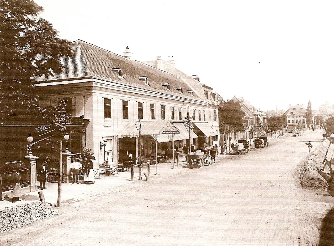 Hietzinger Hauptstraße 10–14 in Wien-Hietzing, 1889. Im Bild das "Dommayersche Kasino", an dessen Stelle 1907 das Parkhotel Schönbrunn errichtet wurde. Gasleuchten als Straßenbeleuchtung, vorne mit Geländer, zum Ablegen von Pferdesattel und Zügel, und Radabweiser. Straße planiert, beidseits liegen Haufen, vermutlich Kleinpflaster zur Verlegung. – Albuminpapier auf Untersatzkarton.