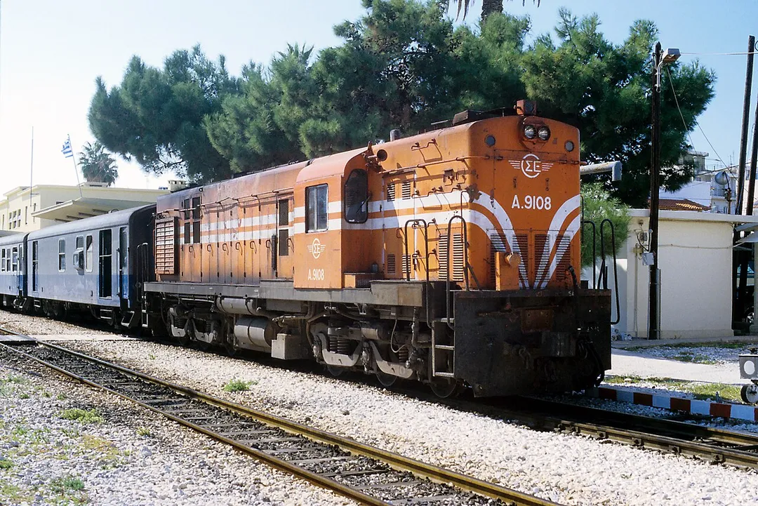 Personenzug 301 Pyrgos–Agii Anargiri, er verlässt Kórinthos 14.35. Der Wagenzug besteht aus Ammendorfer Wagen, erkennbar an den Übersetzfenstern in UIC-Bauart, den glatten Dächern und nach innen öffnenden Türen. Die Lokomotiven der Reihe A.9101 sind typische Alco-Konstruktionen, erkennbar an der Bauart der Schwanenhalsdrehgestelle und der Führerstandsform. Der Nachteil der Bauart ist die schlechte Streckensicht, die in beiden Fahrtrichtungen eine zweimännige Besetzung erfordert. Das Regelspurpendant bei der OSE ist die Reihe A.321 (ALCo DL543), in dieselbe Bauart gehören auch die Renfe-313 (ALCO RSD-39].