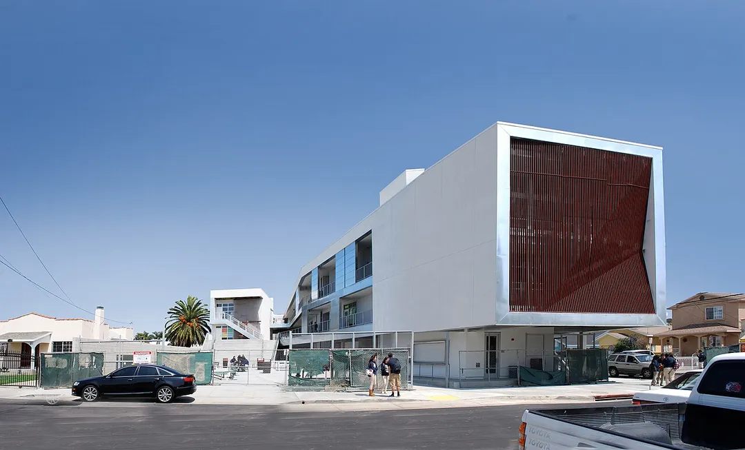 The greatest challenge for the new Animo Leadership High School was posed by the site: undersized and split into two parcels separated diagonally across an intersection. Compounding these issues is the school’s location, near Los Angeles International Airport and within a quarter mile of the 105 Freeway—adding noise and air quality to the list of concerns. The parcels’ small size and disjointed nature made the school’s organization a paramount priority. The solution was to concentrate the programming with the most daily activity on one site—grouping classrooms, administrative offices, and support facilities such as the library and laboratories—leaving the other site to accommodate large assembly spaces that incur less frequent traffic, in this case the multipurpose gymnasium and a large studio space for music and dance.
With a project target of CHPS and/or LEED Certification at the minimum, aesthetics, sustainability, and cost-effectiveness were considered in every design decision. The south facade is clad with 650 solar panels. When completed the project is expected to reduce carbon emissions by over 3 million pounds.
Taking full advantage of the region’s temperate climate, the designers eschewed the fully contained “big box” idiom of conventional schools on the primary use site. Instead, a landscaped courtyard with multifunctional “bleacher” terracing flows into the open-air covered lobby and the multilayered paseo, lending the school the appeal of a collegiate campus and offering significant environmental benefits—improving daylighting and access to fresh air both inside and out—while providing substantial cost savings by limiting artificial lighting and thermal conditioning to the smaller enclosed spaces.

Faceted, ribbed screens appear as a façade motif; both visually connecting the school with the community and modulating light to protect from the building from unwanted solar heat gain. Solar panels form the screen system on the longest façade of the building; augmented by additional panels on the roof, they will provide clean, renewable energy for the school. On the other site, the multipurpose gymnasium and music/dance studio are housed on either end of a long 35’-high building with shared support space in between. Glazed 10’-high roll-up doors that spread across both ends maximize daylighting and views, and can be opened to extend activity into the courtyards beyond. Numerous other sustainable design features and environmentally-friendly materials are incorporated throughout the entire project.