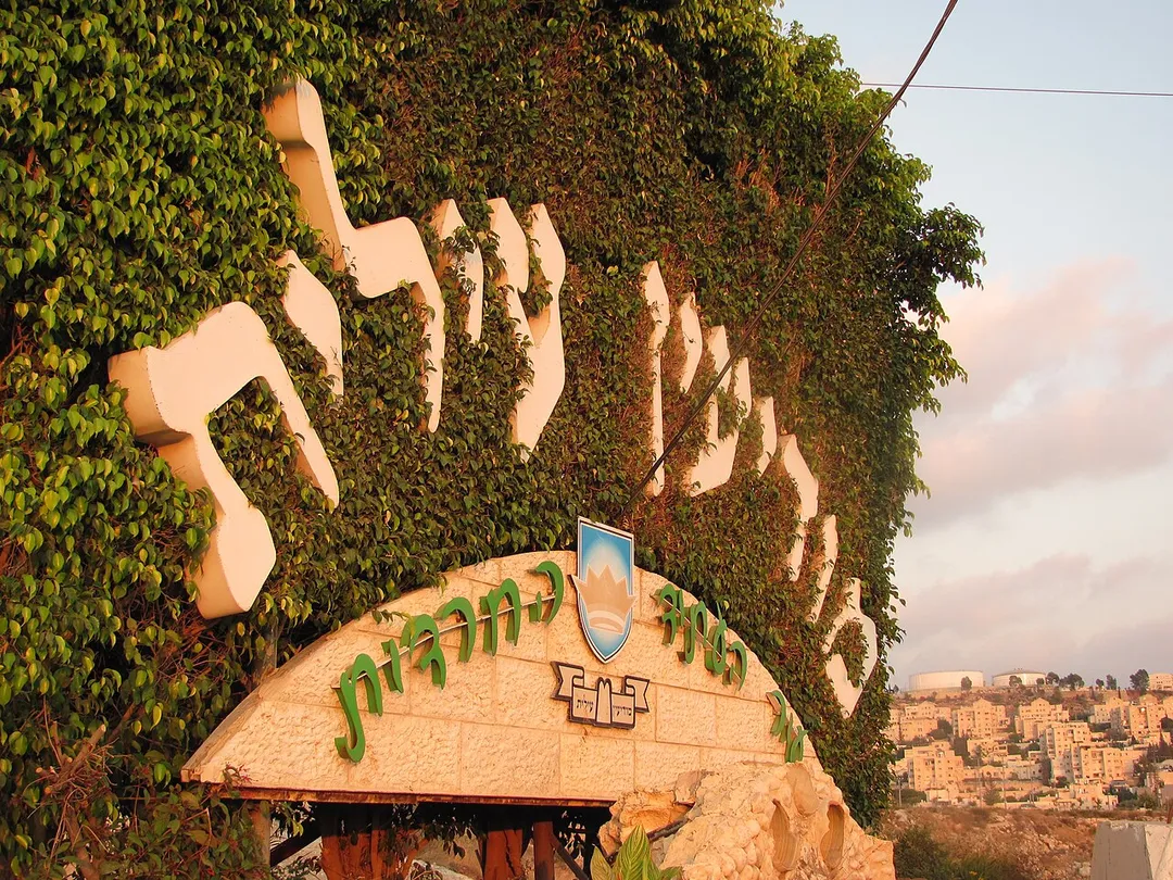 Welcome sign outside the entrance to Modi'in Illit, with homes of Kiryat Sefer in the background. The words "Modi'in Illit" are written in large Hebrew letters on a background of grass. The wording on the stone section of sign reads: "Haredi City of the Future".