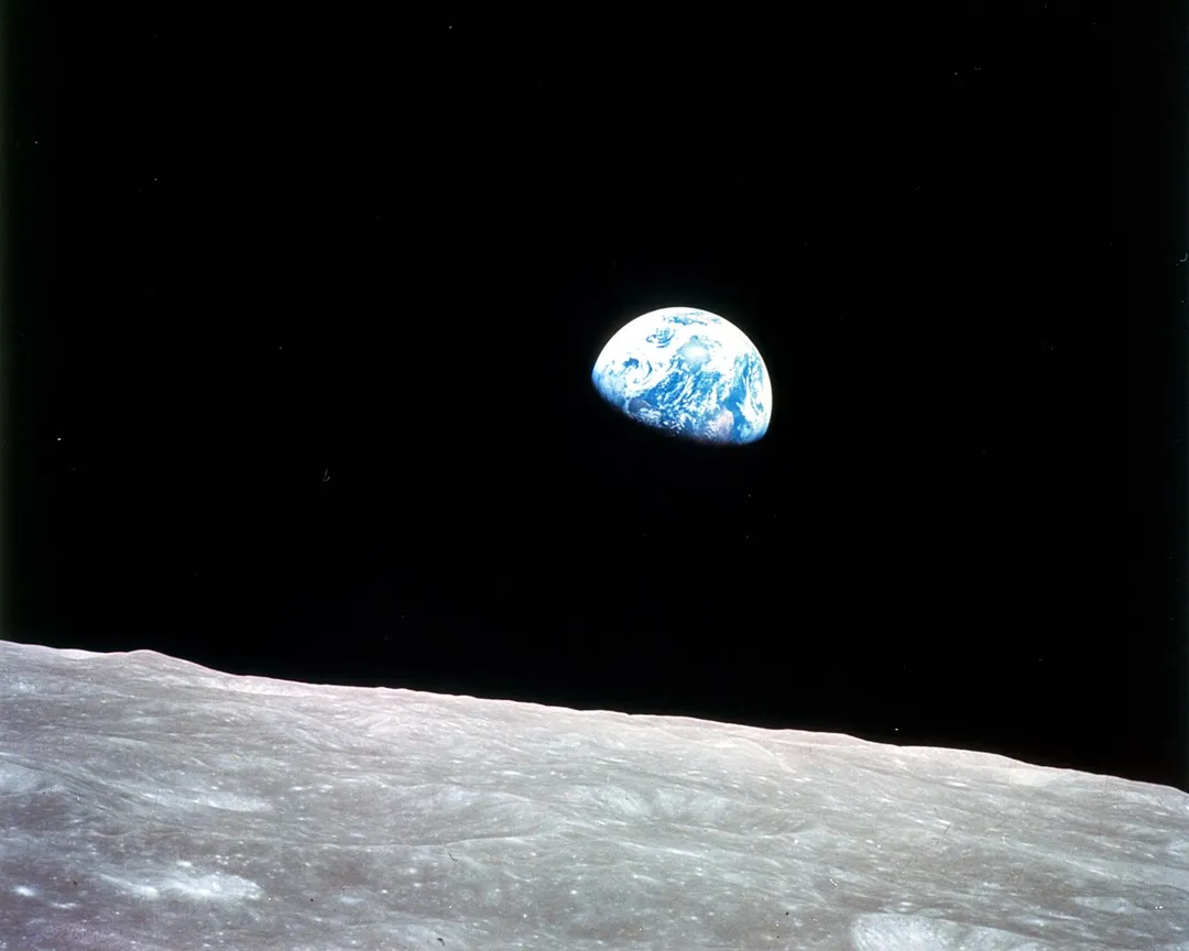 This view of the rising Earth greeted the Apollo 8 astronauts as they came from behind the Moon after the fourth nearside orbit. Earth is about five degrees above the horizon in the photo. The unnamed surface features in the foreground are near the eastern limb of the Moon as viewed from Earth. The lunar horizon is approximately 780 kilometers from the spacecraft. Width of the photographed area at the horizon is about 175 kilometers. On the Earth 240,000 miles away, the sunset terminator bisects Africa.