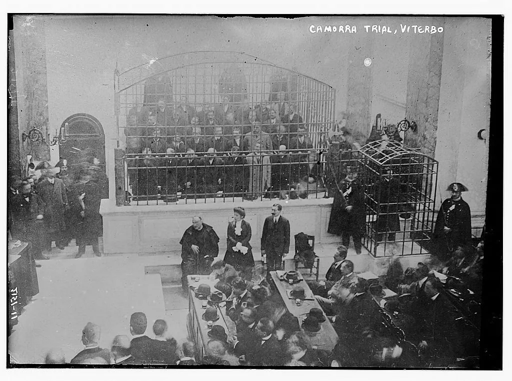 The Cuocolo trial in Viterbo (1911-1912). Most of the defendants are in the large cage. The three in front are (from left to right) the priest Ciro Vitozzi, Maria Stendardo, the only female defendant, and w:Enrico Alfano. In the small cage to the right is the crown witness Gennaro Abbatemaggio. For the caption, see Camorra Verdict; All Found Guilty, New York Tribune, July 9, 1912, p. 4