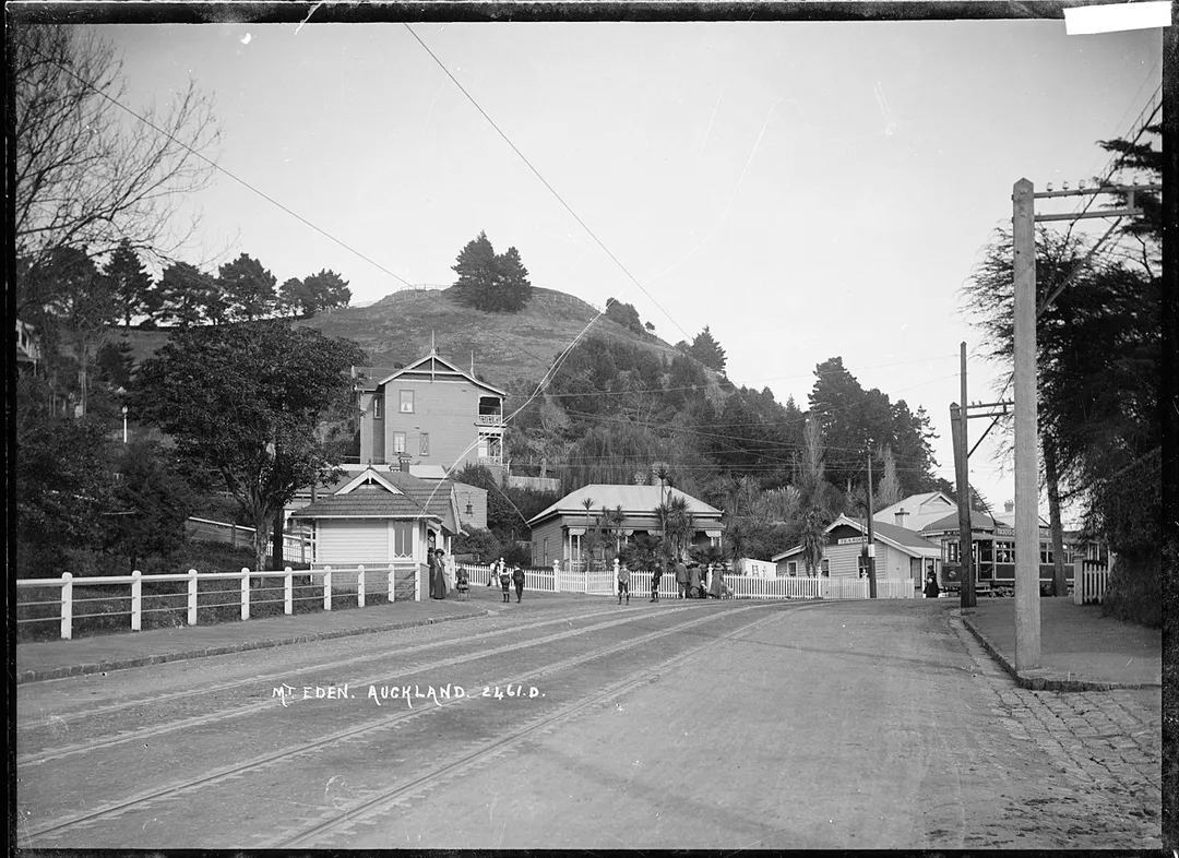 View taken from the corner of Mount Eden Road and View Road looking towawrds the lower slopes of Mount Eden. On the corner of the road leading up to the top of Mount Eden there is a tram stop with people waiting for the tram to arrive. The tram is just visible on the righthand side. A tearoom can be seen in the middle distance on Mount Eden Road. Photograph taken by William A Price in early 1900s.
Inscriptions: Inscribed - Photographer's title on negative -bottom left: Mt. Eden. Auckland. 2461D.
Quantity: 1 b&amp;w original negative(s).
Physical Description: Dry plate glass negative 4.5 x 6.5 inches 

 Mount Eden Road, Mount Eden, Auckland. Price, William Archer, 1866-1948 :Collection of post card negatives. Ref: 1/2-000684-G. Alexander Turnbull Library, Wellington, New Zealand. natlib.govt.nz/records/22744595