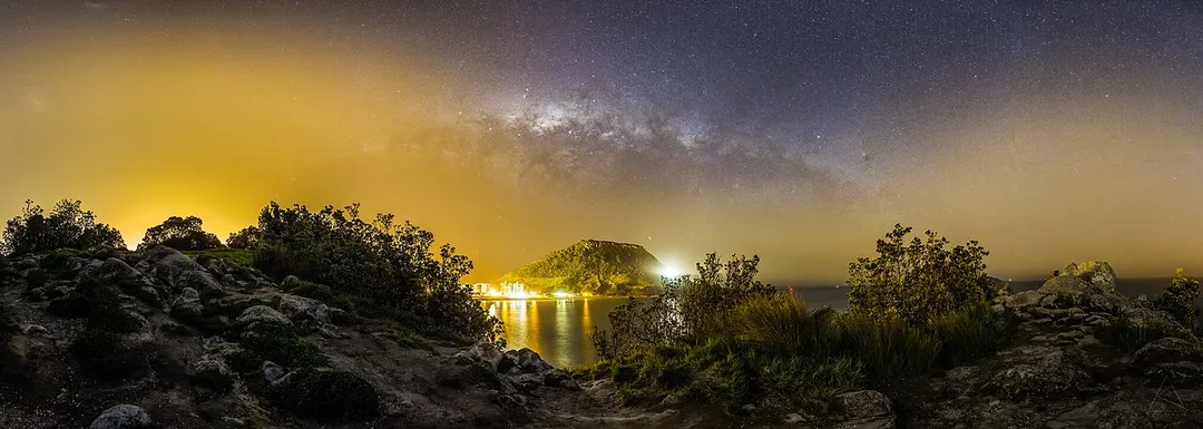 Mauao (Caught by the dawn) waits there for the people of the night to help him fulfill his desire. Rays of the sun sent back the people of night to the depths of forest and looks like they are never coming back because of the light pollution around it.
This images shows how we are losing the beauty of night sky to unshilded artificial lightning. The lights that are supposed to light up the ground is also lighting up the sky.
The stars in the sky around towns and cities are losing their battle to artificial lightning, if nothing is done soon, they will lose it and the future generation will never get to see the night sky and "Twinkle Twinkle Little Star" will be a poem of the past.
Despite the light pollution and sea mist (which enhances the light pollution), was pretty impressed to see the milky-way going across and the Magellan clouds (Two small smudges on left top of the image)

This is a 21 image panorama, stitched together in Lightroom and processed in Photoshop, processing against the light pollution is really tough.