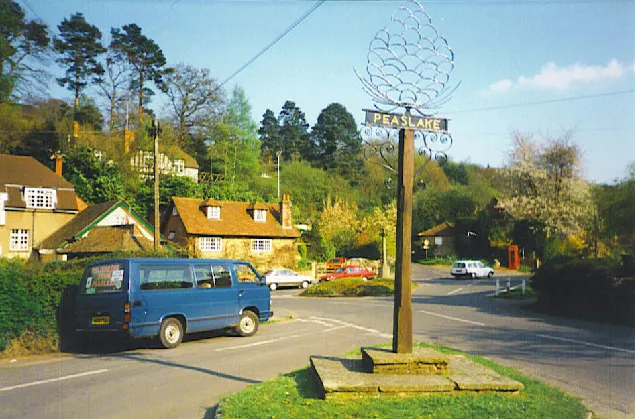 Peaslake, Village Centre. Four roads converge by the village shop and hotel at this crossroads in the Surrey Hills. The village sign is topped by a stylized fir cone, reminiscent of the trees on the surrounding hills.