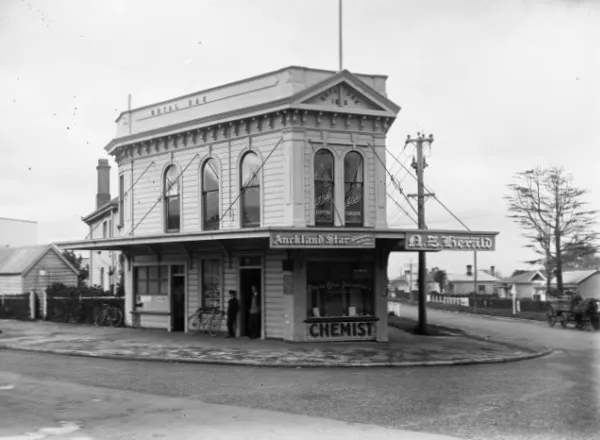 The pharmacy building at what is today the Royal Oak Roundabout in 1910, Royal Oak, Auckland, New Zealand. Looking southeast with Campbell Road to the left, and Mt Smart Road to the right. 

Extended information on origin webpage reads: Business premises on the corner of Campbell Road and Mt Smart Road, Royal Oak, Auckland [ca 1910] / Reference number: 1/2-001197-G / 1 b&amp;w original negative(s). Dry plate glass negative 4.5 x 6.5 inches. Horizontal image. / Part of  Price, William Archer, d 1948 :Collection of post card negatives (PAColl-3057) Photographic Archive / Scope and contents: View of the building on the corner of Mt Smart Road and Campbell Road, Royal Oak. The Royal Oak Pharmacy and a post office occupy the downstairs areas and J E Butler, dental surgeon, has rooms upstairs. Two men can be seen in a doorway (one is a postman) and there are two bicycles nearby. A house and cart can be seen on the road to the right. Auckland Star and NZ Herald signs can be seen. The building at the back with a chimney is the original Royal Oak hotel built in 1853. Photograph taken by William A Price in early 1900s. / Historical notes: William Archer Price lived and practised in Queen Street, Northcote, 1909-1910. / Source: New Zealand Post Office directories