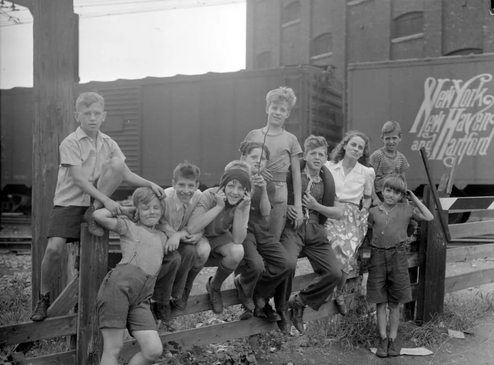 We see the woman of letters Gabrielle Roy surrounded by nine children (eight boys, one girl; Yvon Laferrière is the first on the left) of the St. Henri neighborhood in Montreal. They sit on a fence at the corner of St. Augustine and Saint-Ambroise near the railroad tracks in front of a car in New York, New Haven and Hartford Railroad.