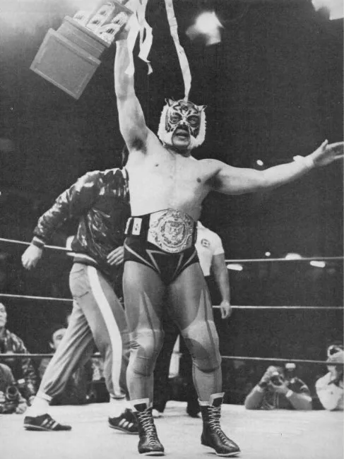 Tiger Mask portant la ceinture de champion poids lourd junior de la World Wrestling Federation ainsi qu'un trophée célébrant sa victoire.