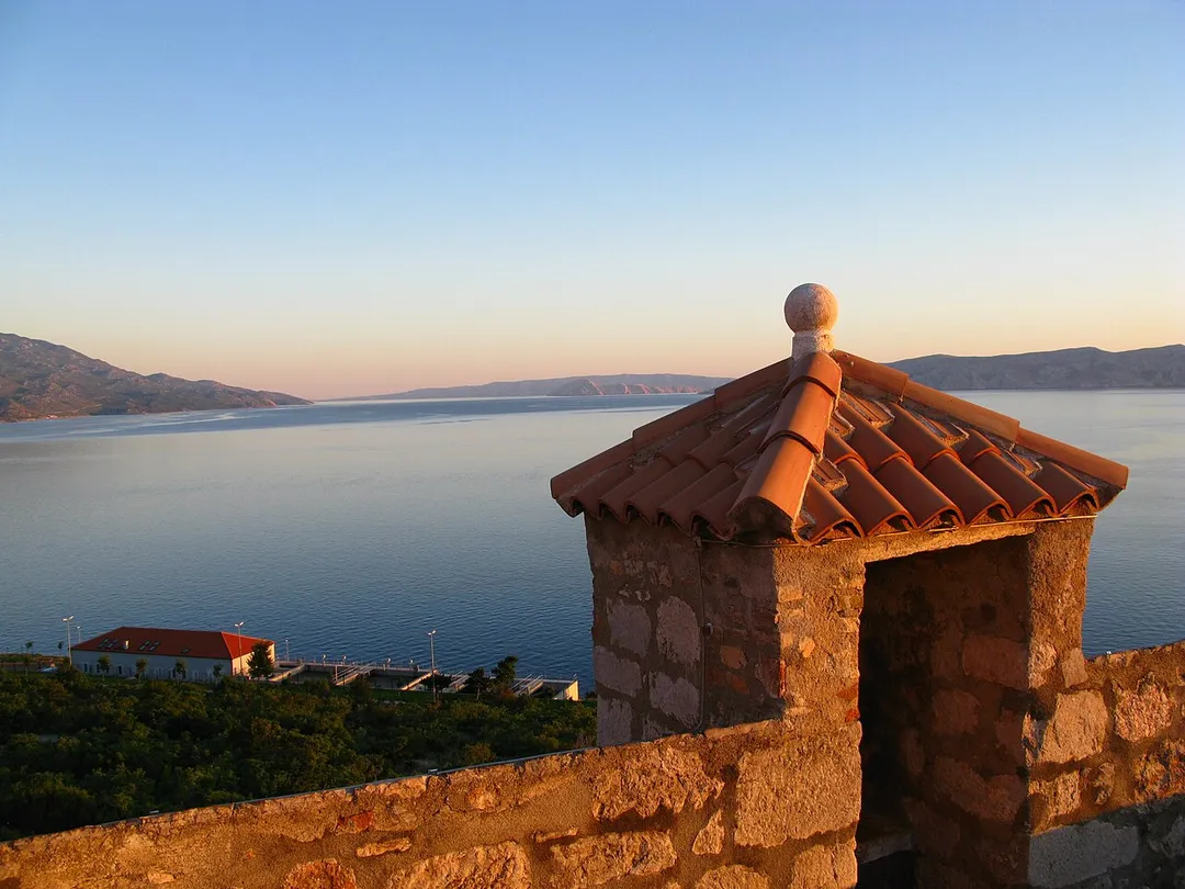 Outhouse of Fortress Nehaj in Senj / Croatia / Adriatic Sea at sunset. Behind the wastewater treatment plant of Senj, on the left side the Velebit-Mountainrange, on the right side the islands of Prvić, Goli Otok and Rab. In the background with the red roof and the pool, the WWTP of Senj, left the Velebit Mountains. The roof of the toilets, where the droppings fall along the outer castle wall in the outdoors, is with the red roof tiles typical of southern Europe (Monk and Nun). The rooftop is adorned with a stone ball.