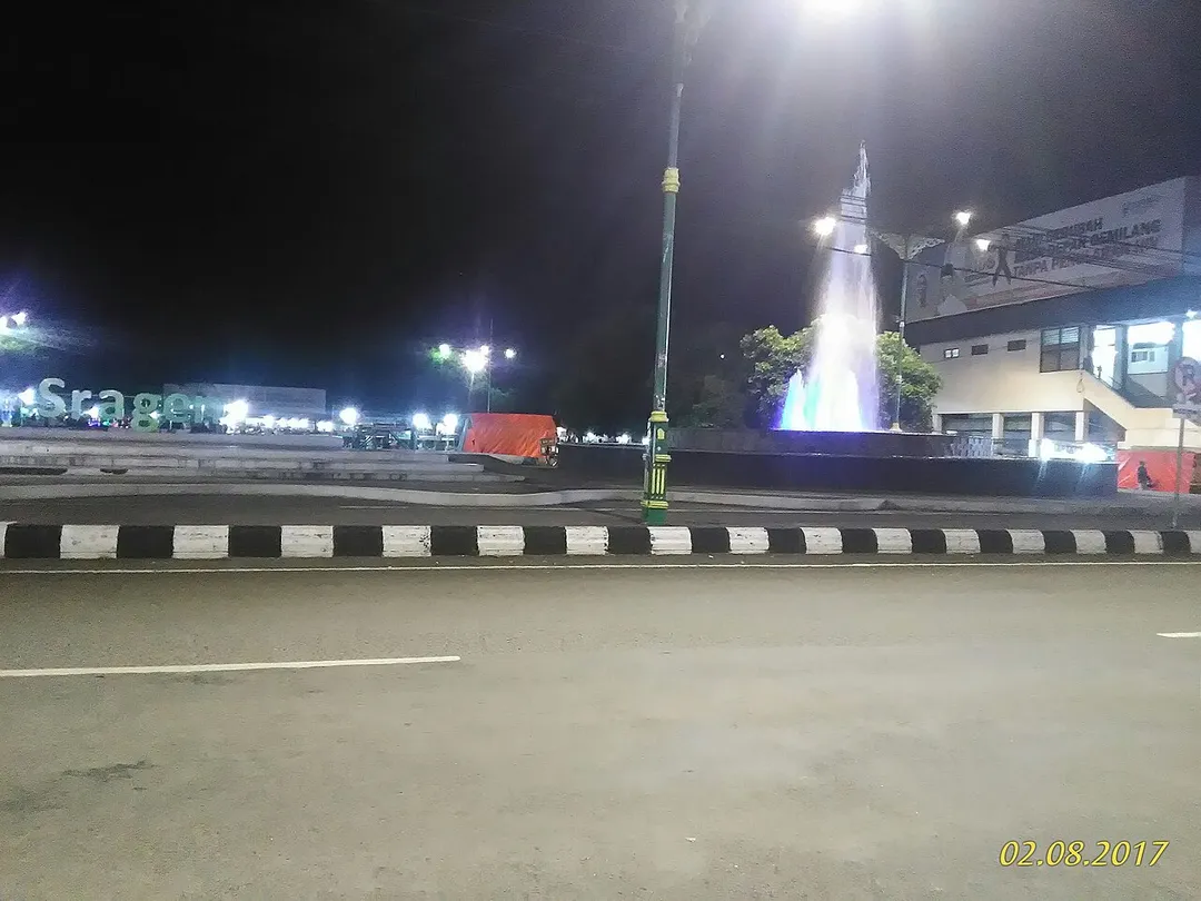Salah satu sisi alun-alun Sragen. Diambil gambar dari sebrang jalan raya, nampak pemandangan air mancur, dan tugu lampu alun alun, serta nama kota yang di buat dari semen dll.