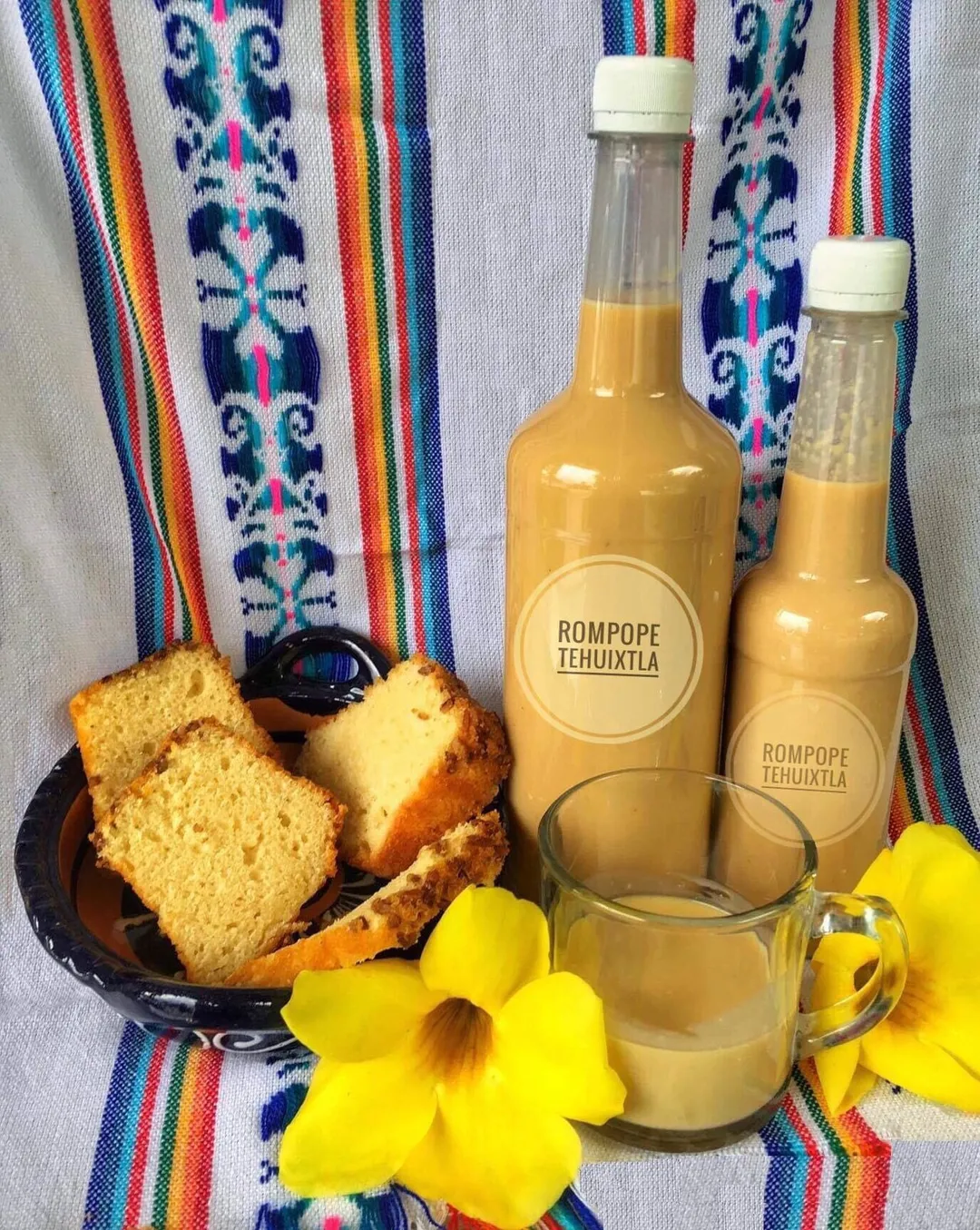 Dos botellas de rompope de Tehuixtla, una taza con el sabroso postre y una cazuelita con rebanadas de pan de nuez con dos flores amarillas y una servilleta de fondo.