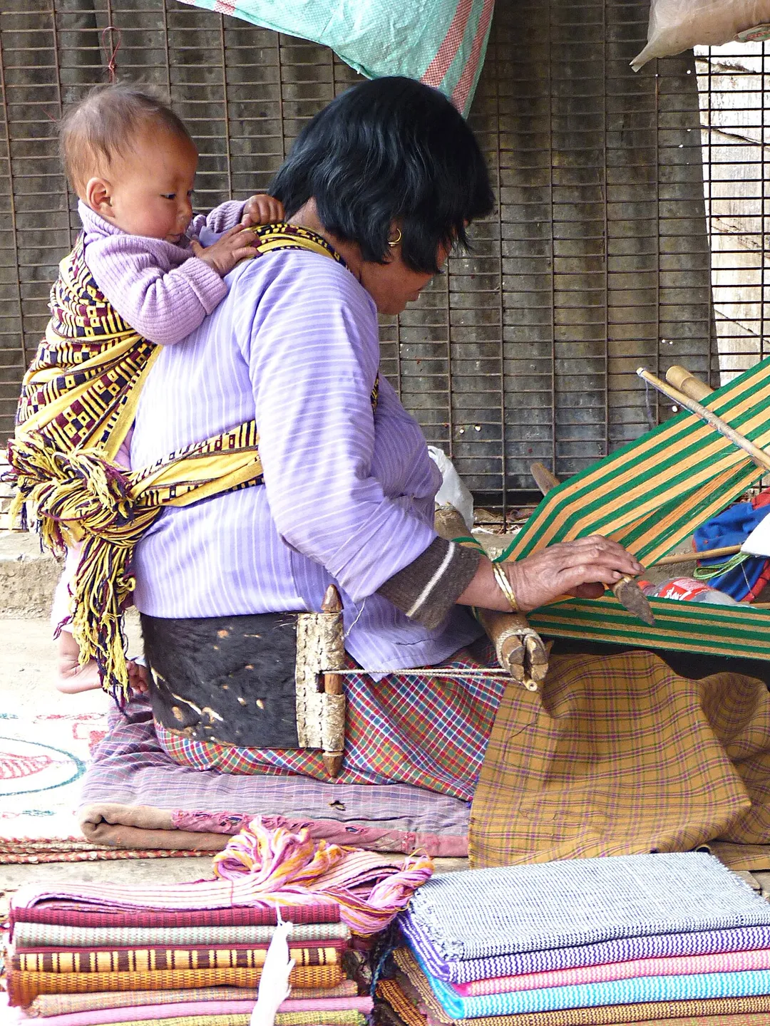 Eine Weberin aus {{:de:Bhutan|Bhutan}} am traditionellen Webstuhl mit Säugling in der Rückentrage, Thimpu